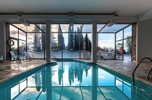 Una piscina interior con paredes de vidrio transparente, suelo de ladrillo y vistas a árboles y edificios exteriores.