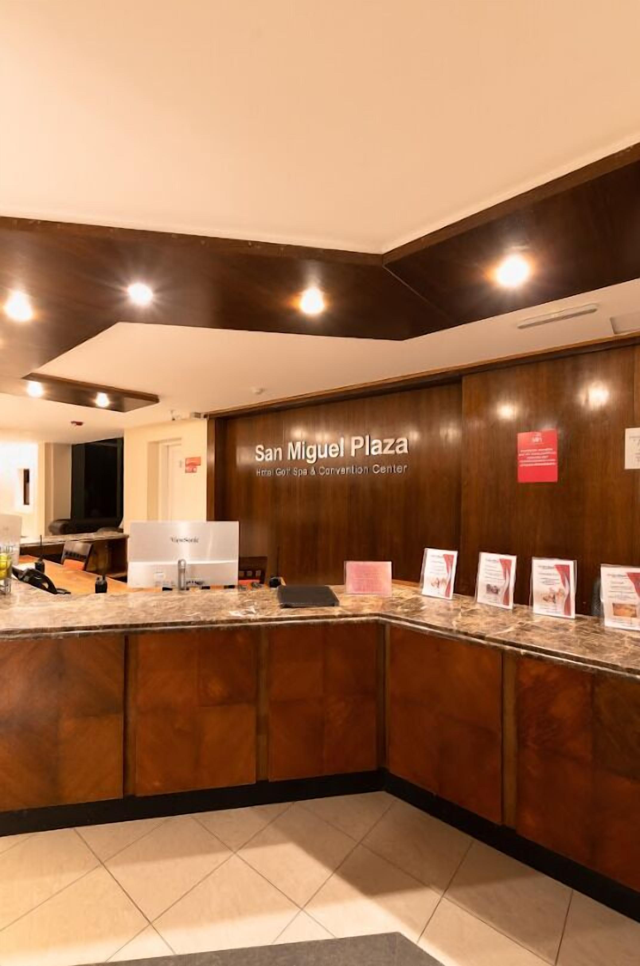 A reception area with a wooden counter, a TV screen, and a sign reading 'San Miguel Plaza.'