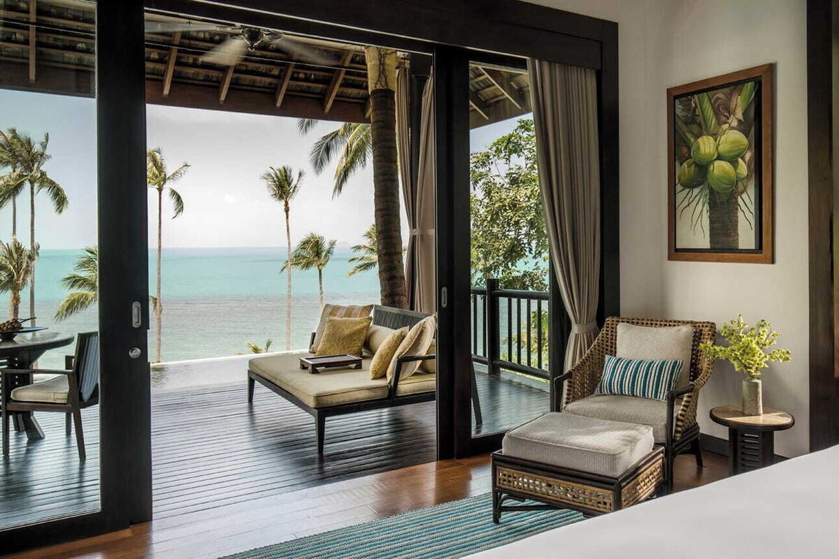 A bedroom with a large bed, a sofa, a chair, and a small table. There is a view of the ocean and palm trees through the open door.