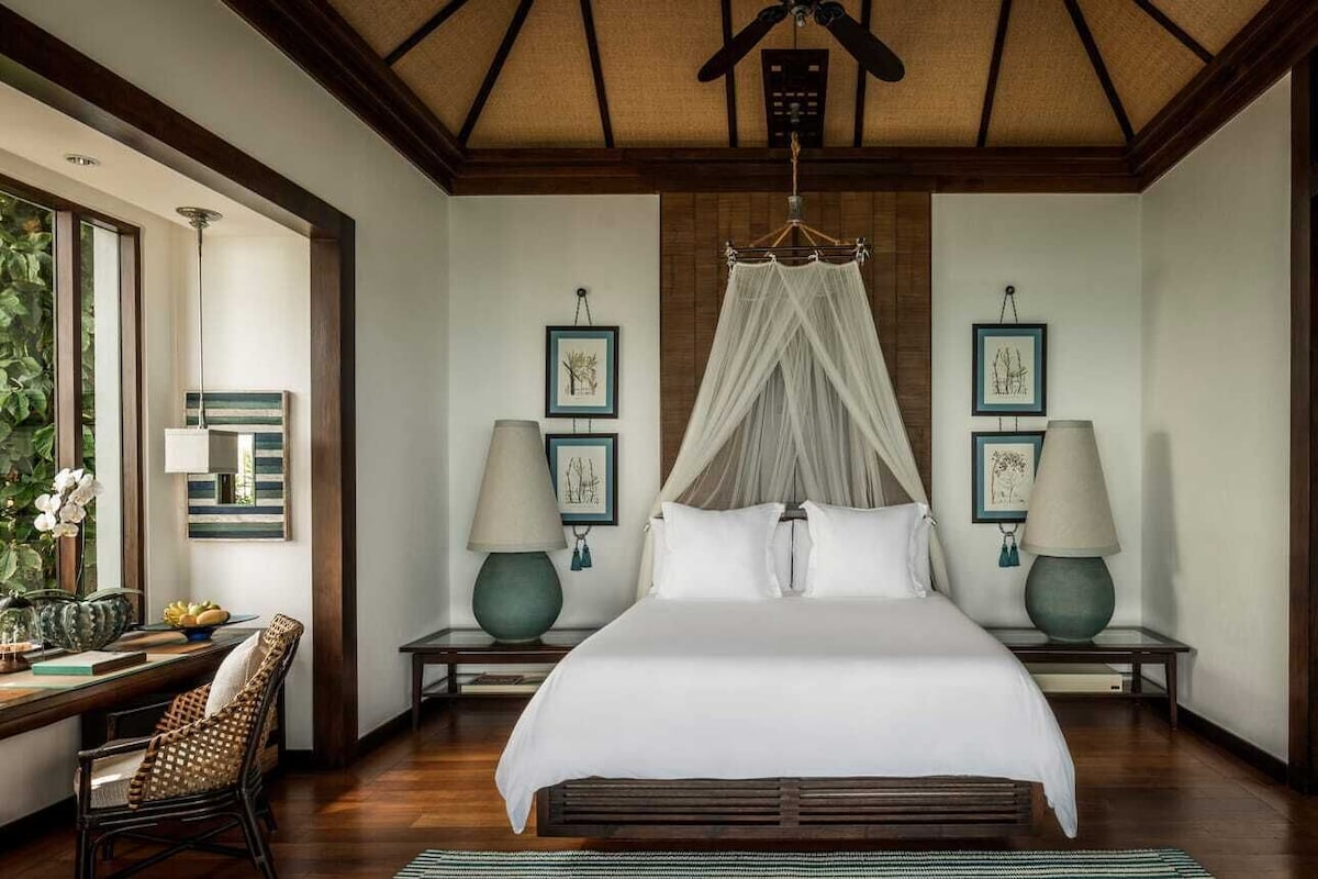 A bedroom with a canopy bed, two bedside tables with lamps, framed artwork on the walls, and a dining area with a chair and table.