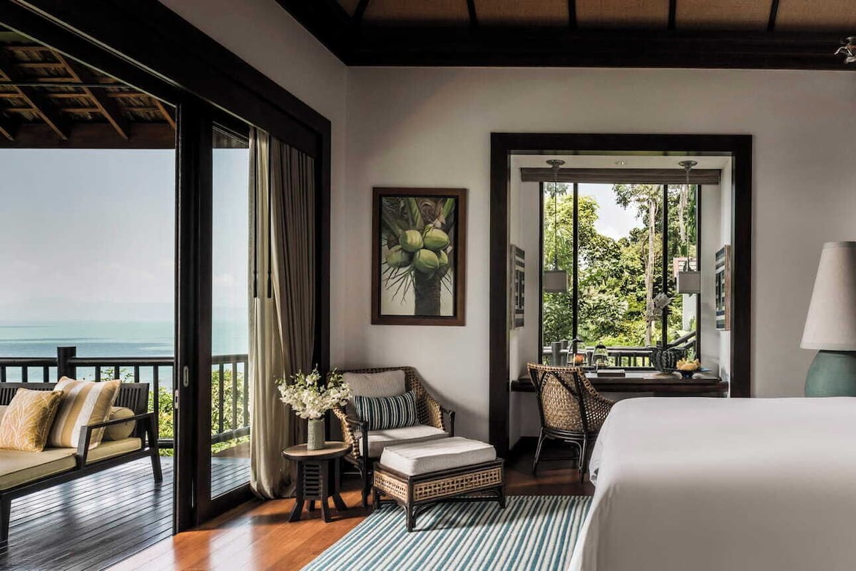 A bedroom with a large bed, a view of the sea, wicker furniture, and a framed picture on the wall.