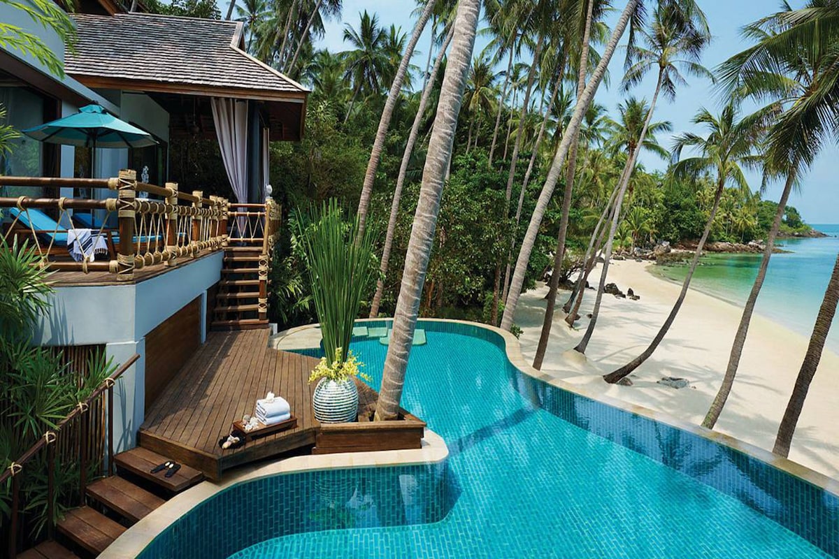 A private pool area with a wooden deck, lounge chairs, and a view of a beach with palm trees.