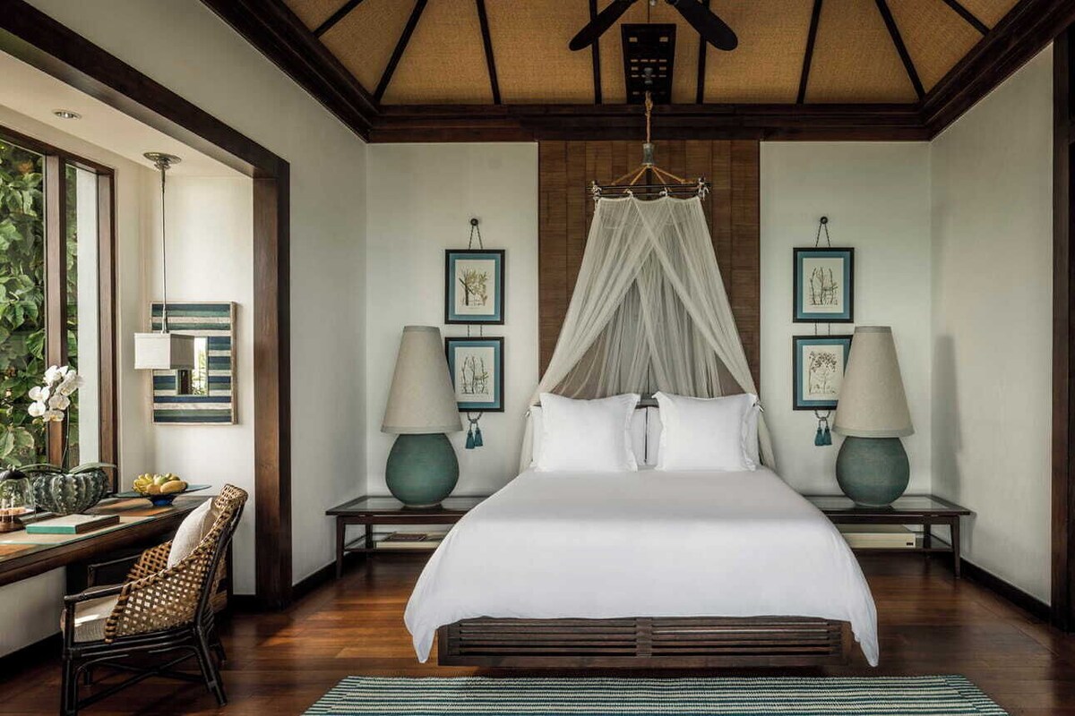 A spacious bedroom with a large bed, two bedside tables with lamps, a canopy bed, framed artwork on the walls, and a view of greenery through a window.