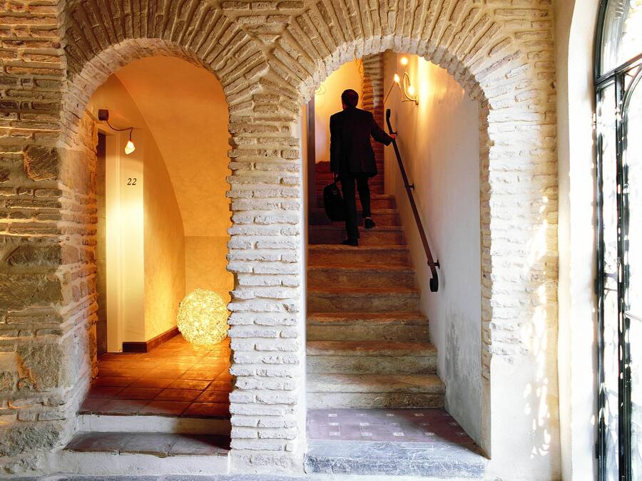 Un escalier en pierre, avec une personne qui le descend, flanqué de murs en briques surmontés d’arcs, et un luminaire sphérique au bas de l’escalier.