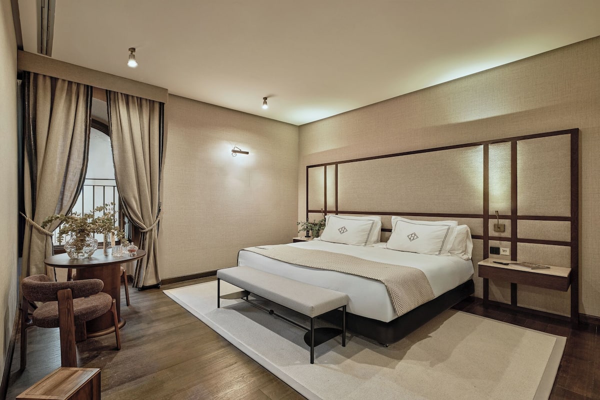 A modern bedroom with a large bed, a bench, a round table with chairs, and a small bedside table.