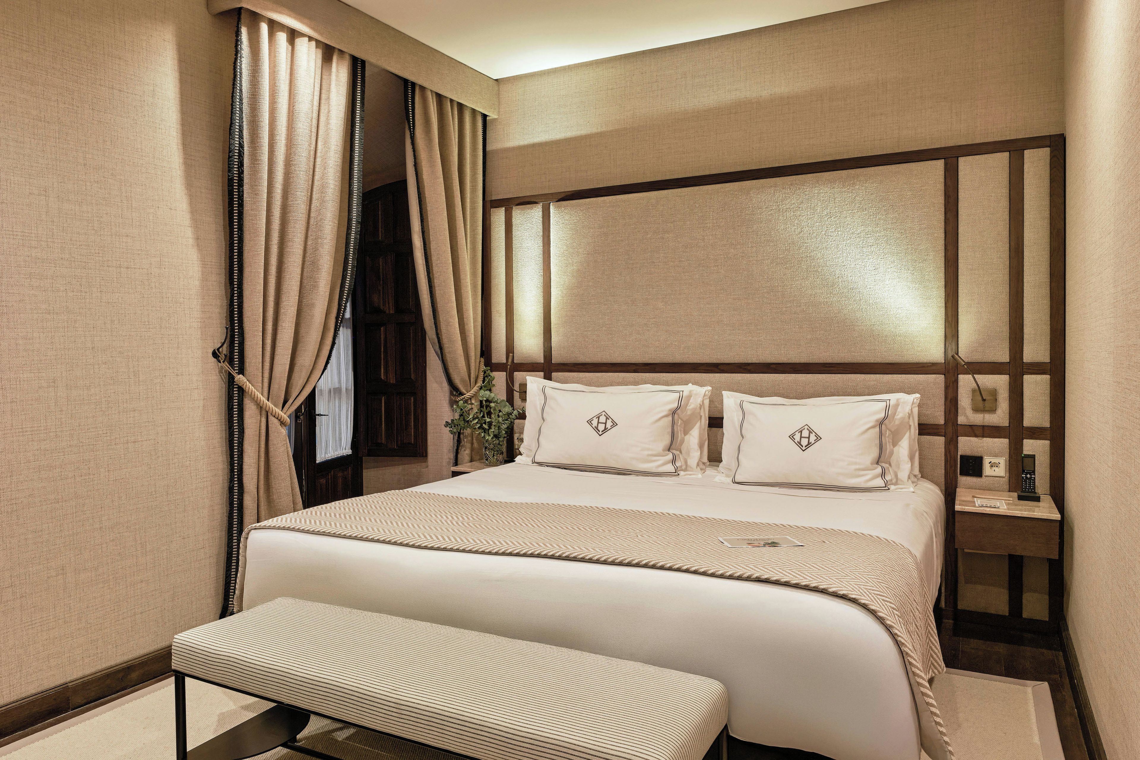 A hotel room with a large bed, a bedside table, a nightstand, a bench, and a window with curtains.