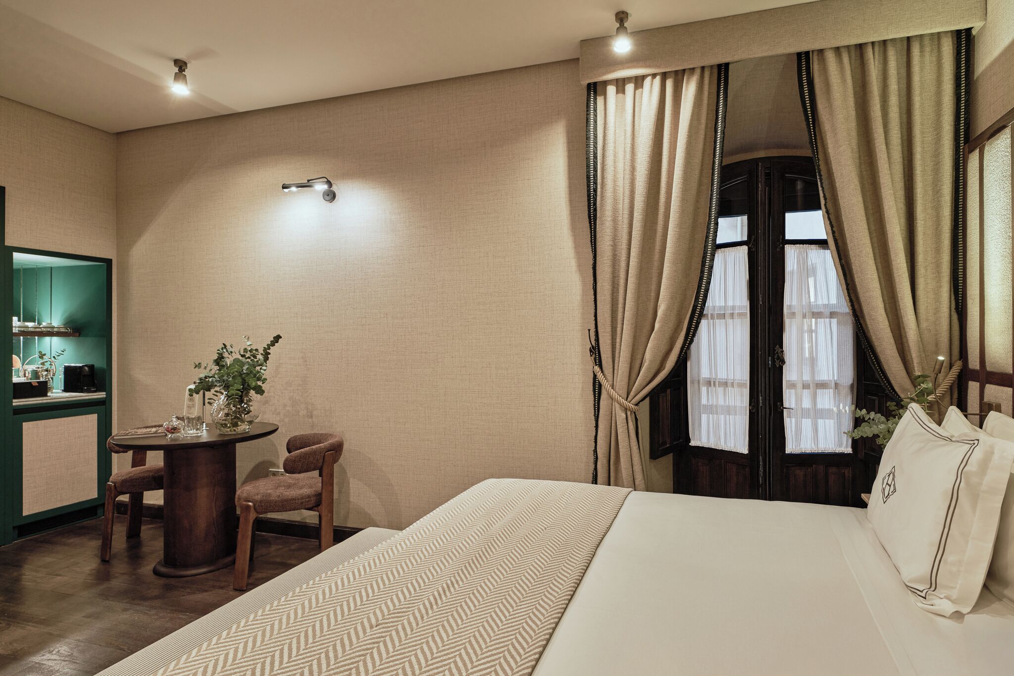 A hotel room with a bed, a small table with chairs, a window with curtains, and a mirror.