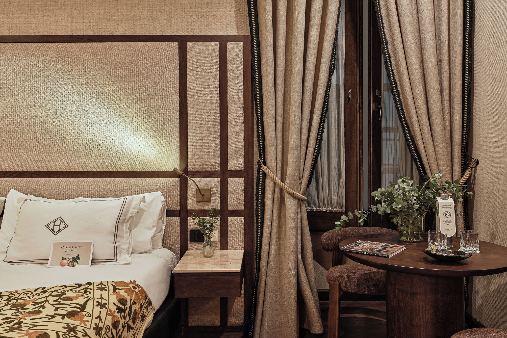 A neatly arranged hotel room with a bed, bedside table, and a small round table with a vase and glasses.