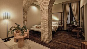 A modern hotel room with a stone archway, a sofa, a small round table with a plant, and a bedside table with a lamp.