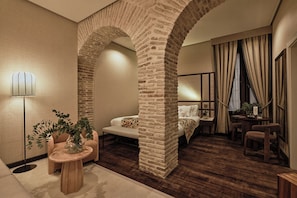 A modern hotel room with a stone archway, a sofa, a small round table with a plant, and a bedside table with a lamp.