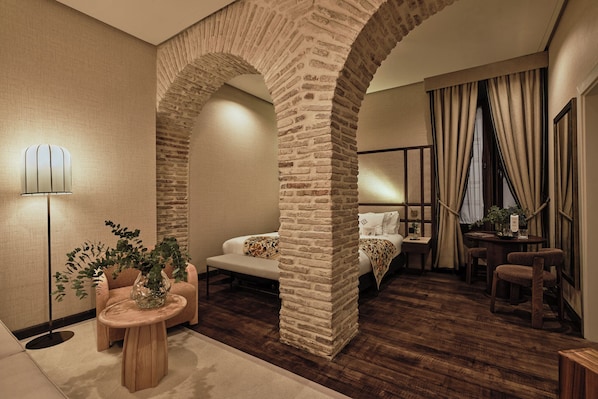 A modern hotel room with a stone archway, a sofa, a small round table with a plant, and a bedside table with a lamp.