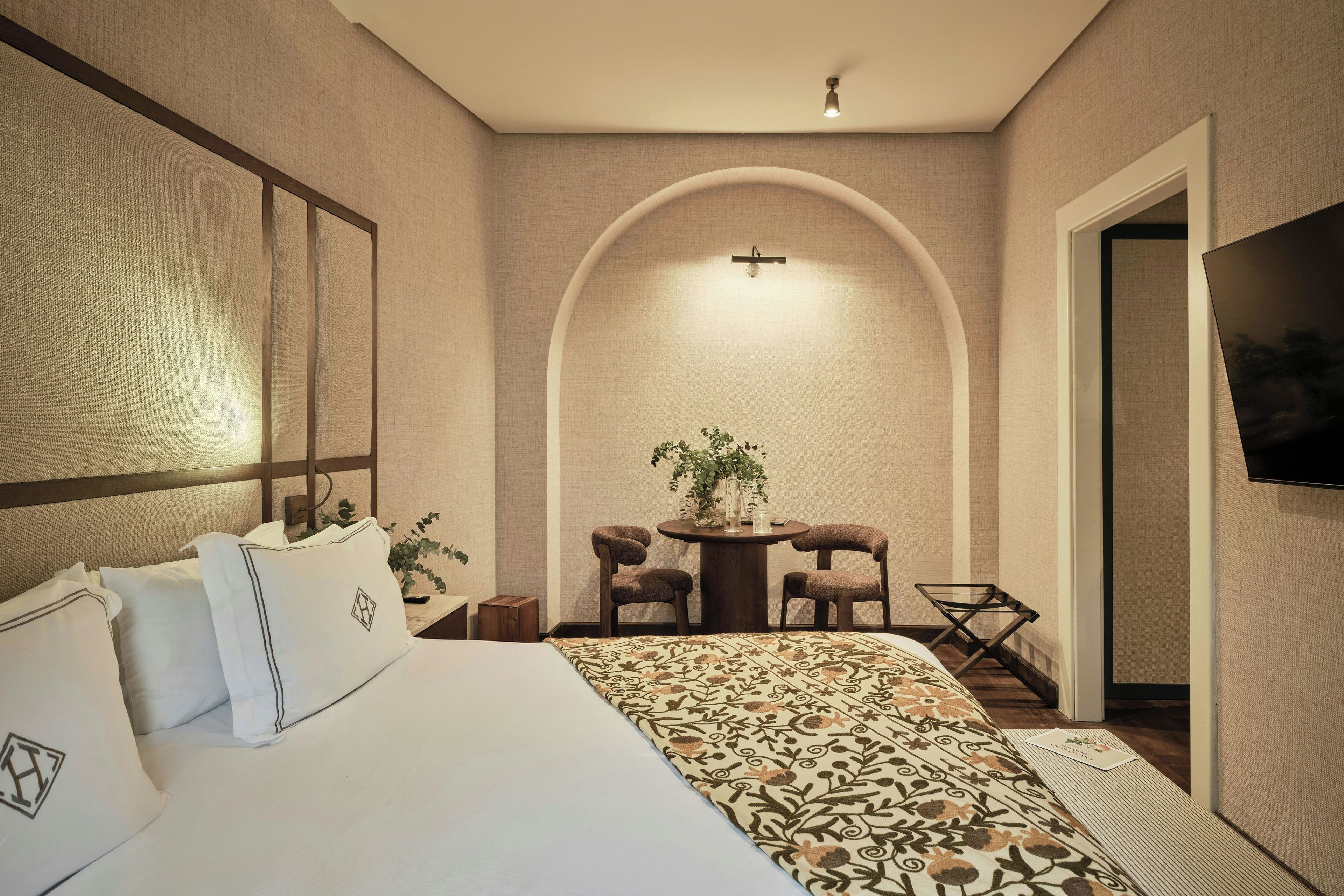 Habitación de hotel con una cama, un escritorio, dos sillas, una mesita con una planta y un televisor en la pared.