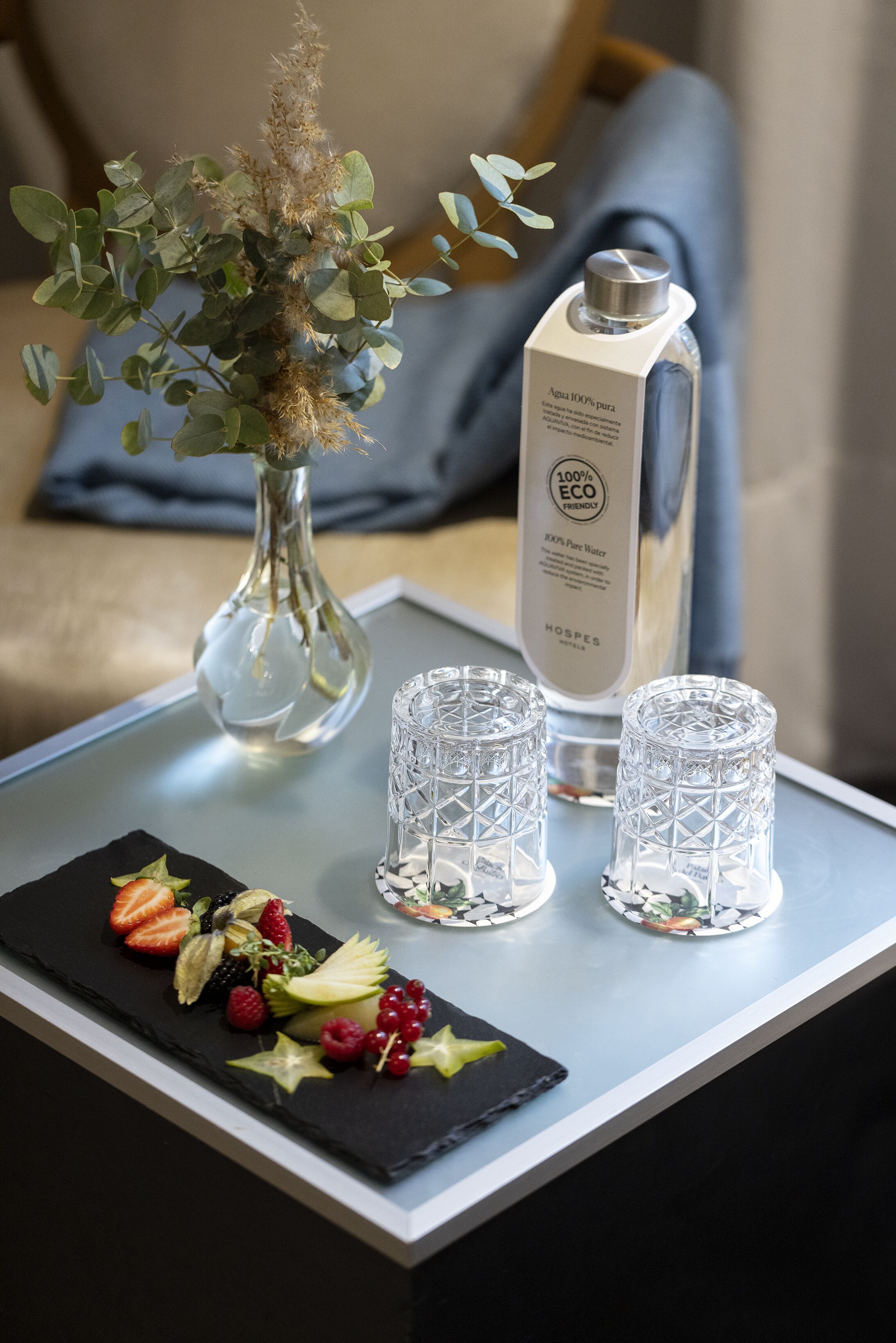 A table with a bottle of water, two glasses, a vase with dried plants, and a fruit platter.