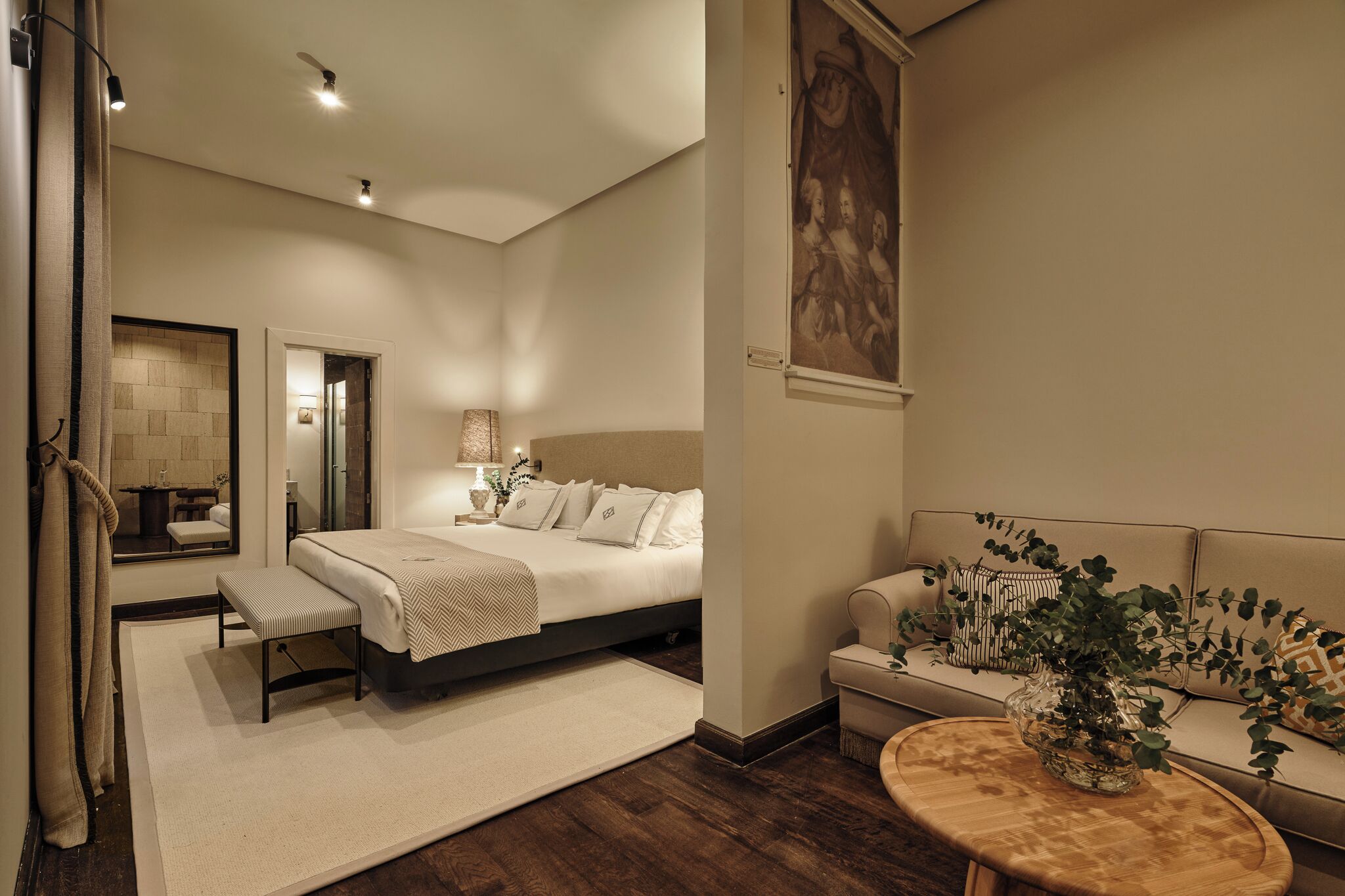 Un dormitorio con cama, mesita de noche, banco y sofá con un jarrón de plantas.