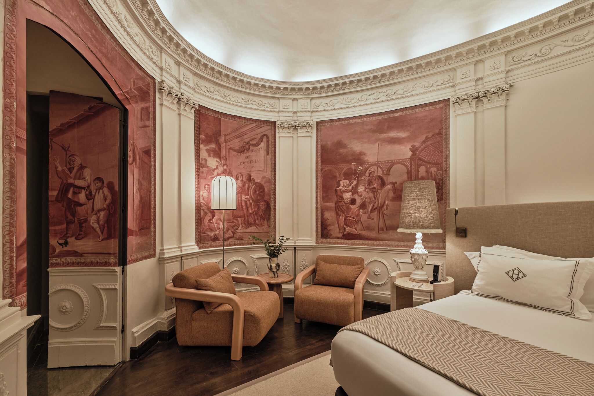 Habitación de hotel con una cama grande, dos sillones, una mesita de noche con lámpara y un mural en la pared.