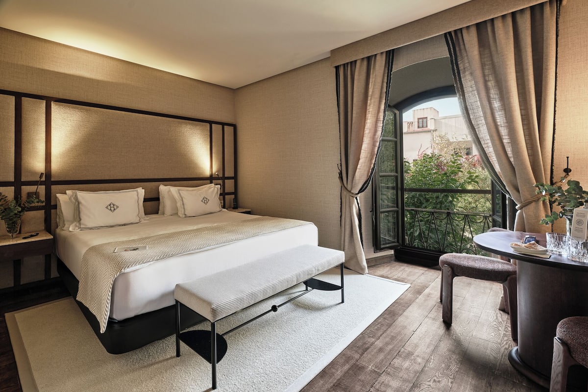 A modern bedroom with a large bed, a bedside table, a bench, a small table with a chair, and a view of greenery through a window.