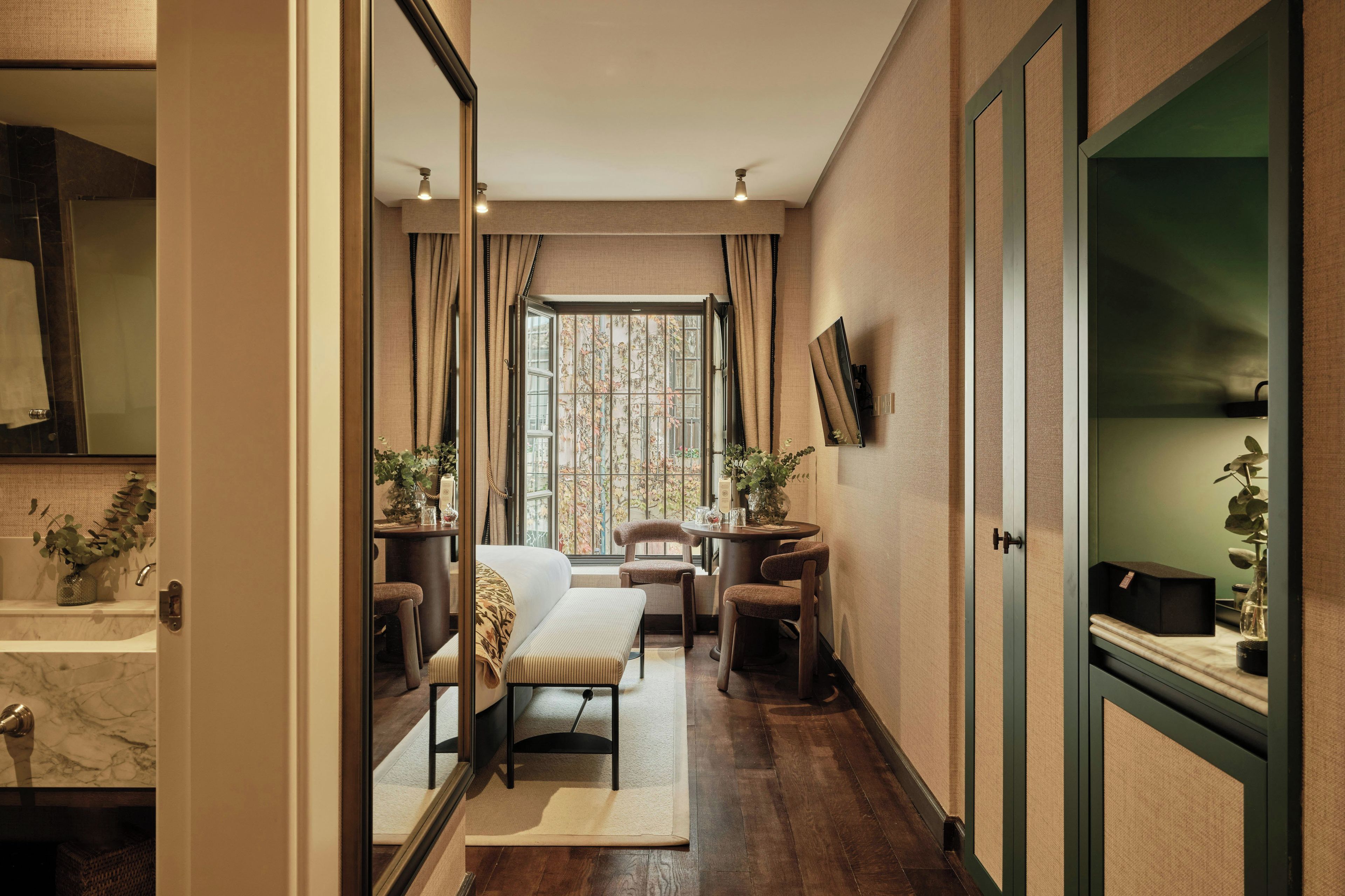 A hotel room with a large window, a sofa, a dining table with chairs, and a mirror on the wall.