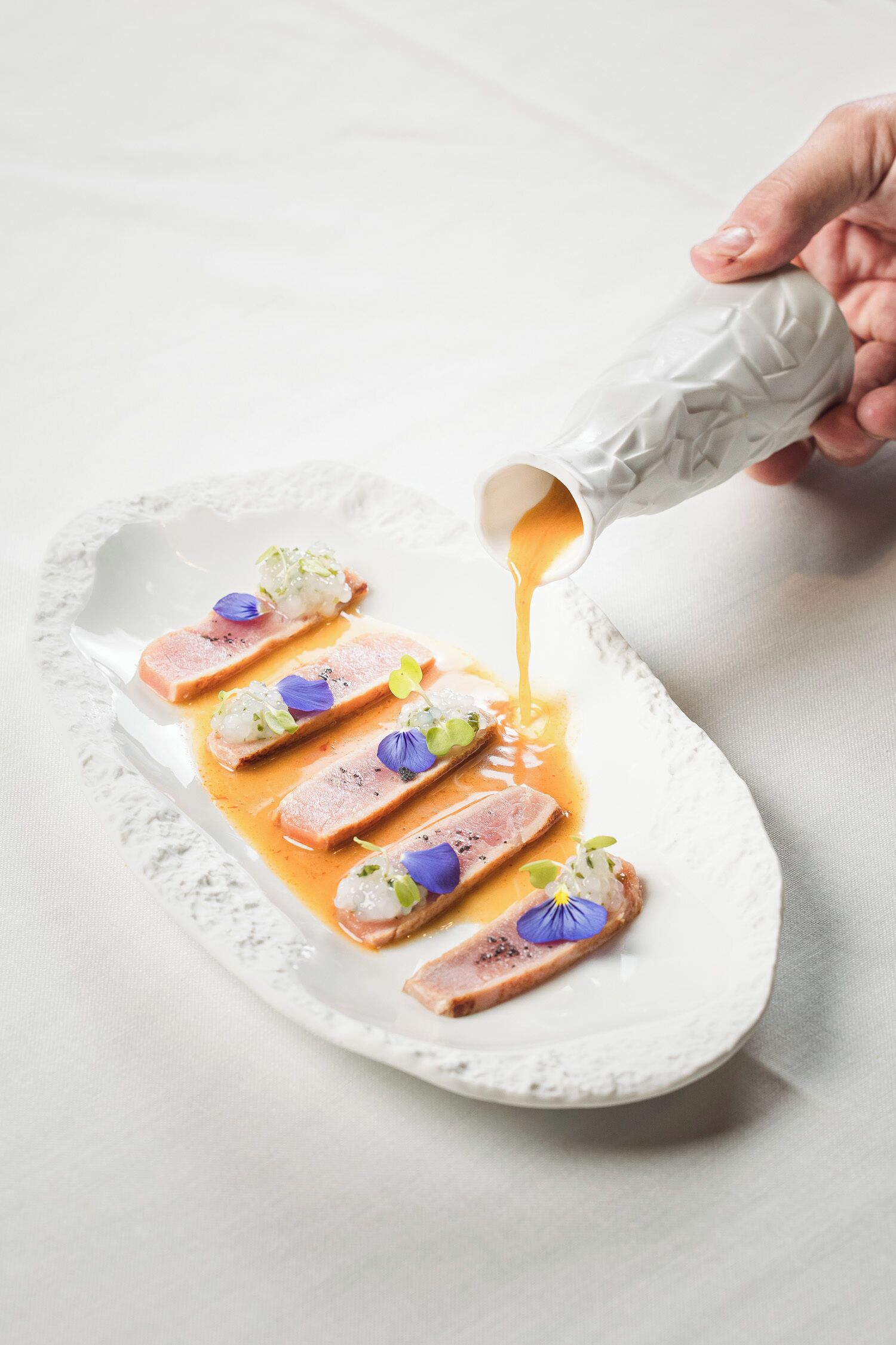 Un plato de cerámica blanca con comida dispuesta en línea, una mano vertiendo salsa sobre él y un mantel blanco al fondo.