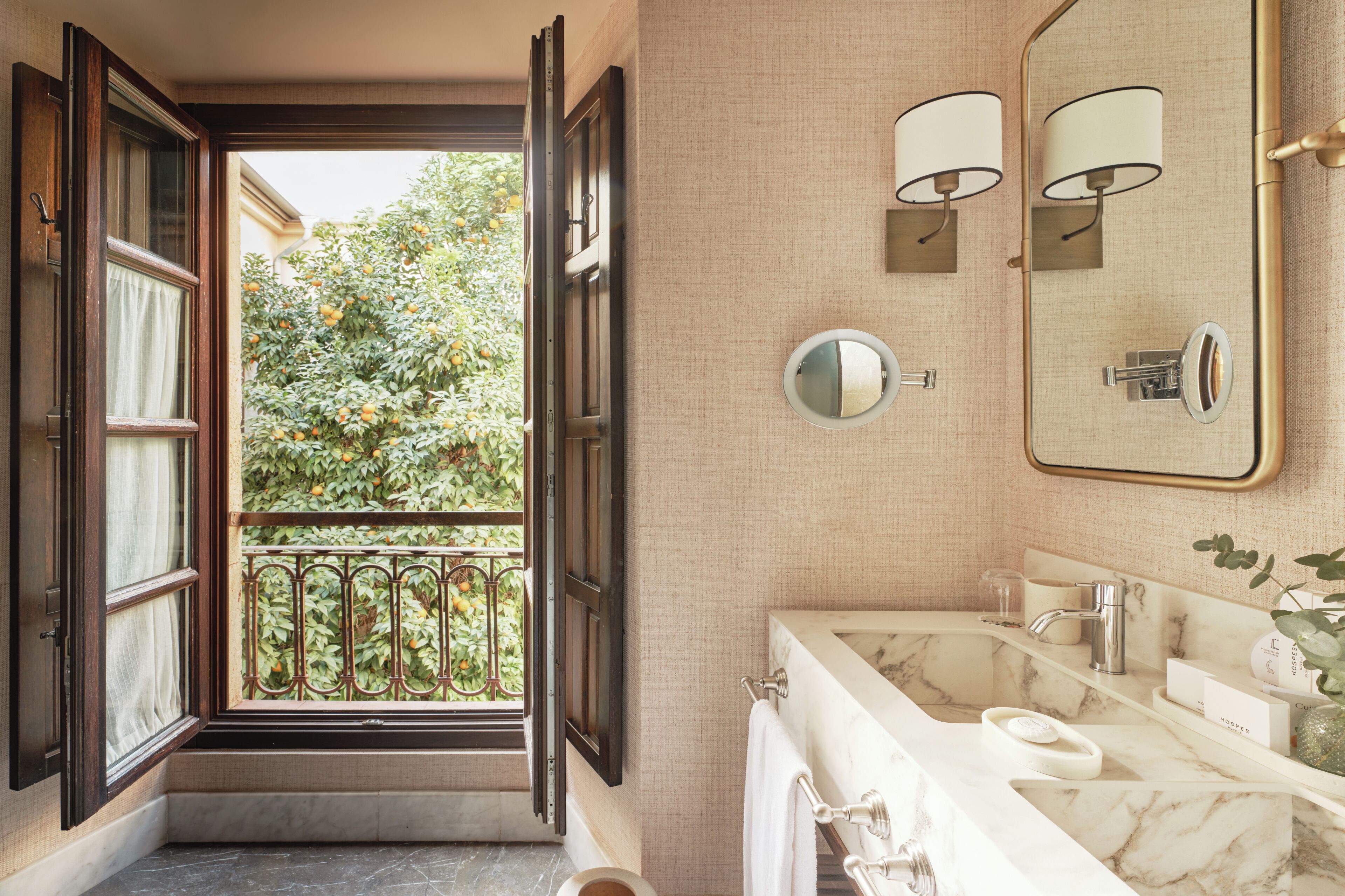 A bathroom with a large mirror, two wall-mounted lights, a marble sink, and a view of a balcony with greenery.