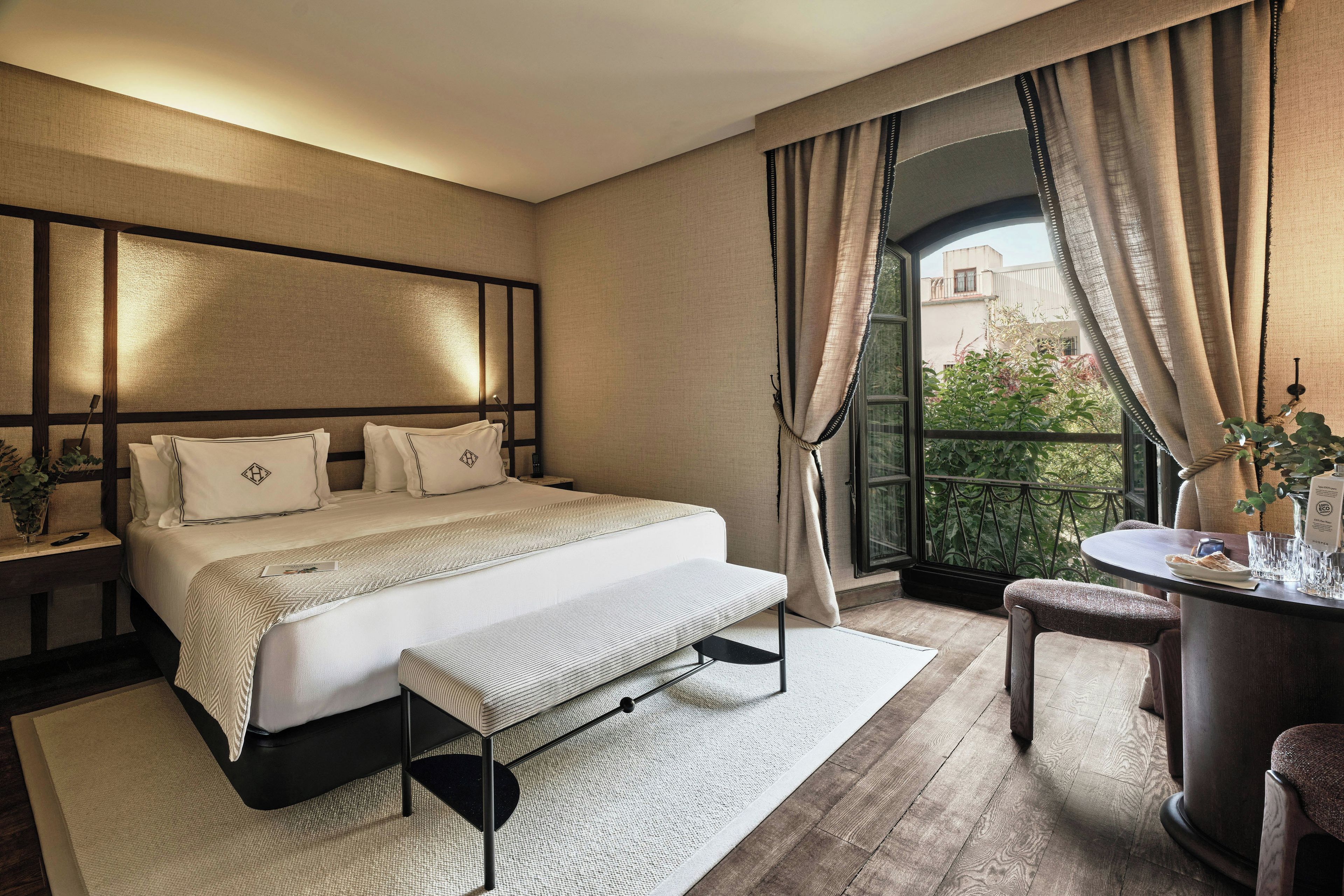 A hotel room with a large bed, a bedside table, a small round table with a chair, and a view of greenery outside.