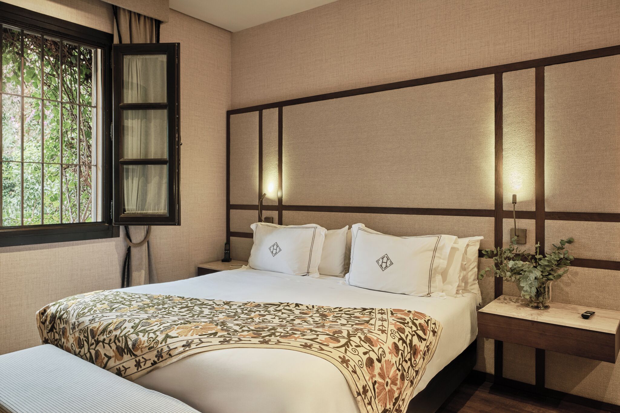A neatly made bed with a patterned bedspread, a bedside table with a vase of flowers, and a window with a curtain.