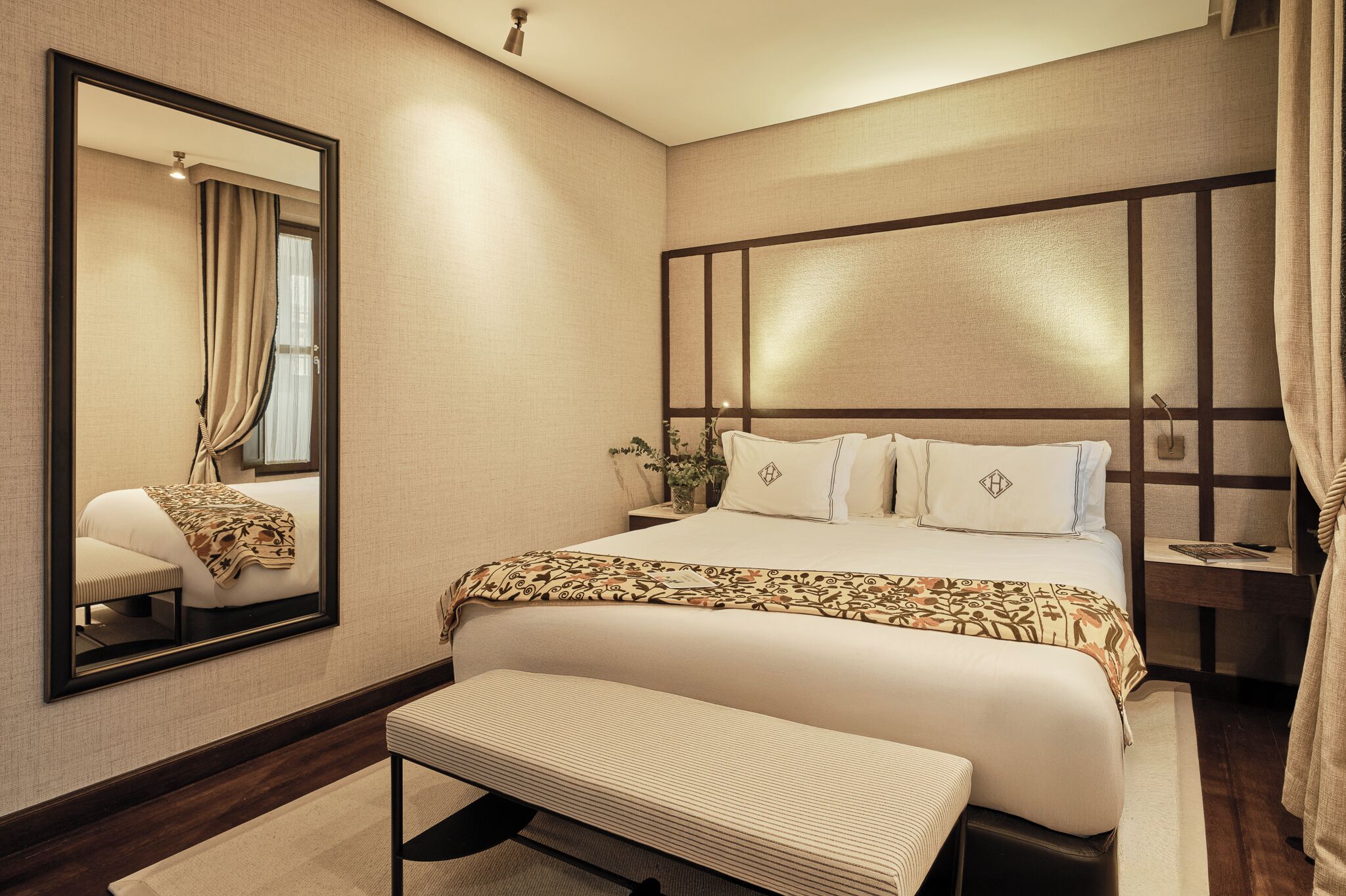 Une chambre d’hôtel moderne avec un grand lit, une table de chevet, un banc, un miroir et une fenêtre avec des rideaux.