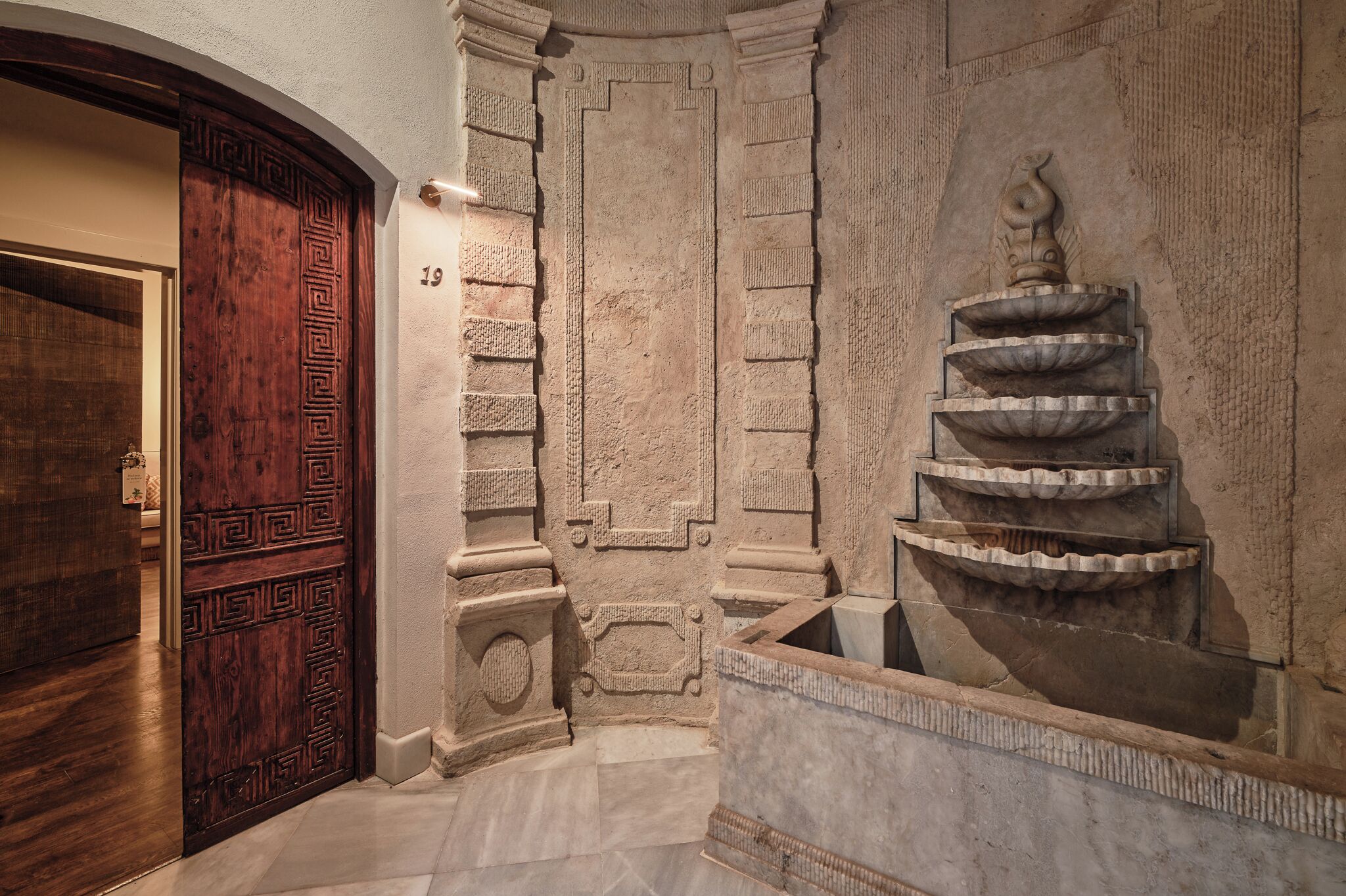 A hallway with a wooden door, stone wall with a decorative fountain, and a number '19' on the wall.