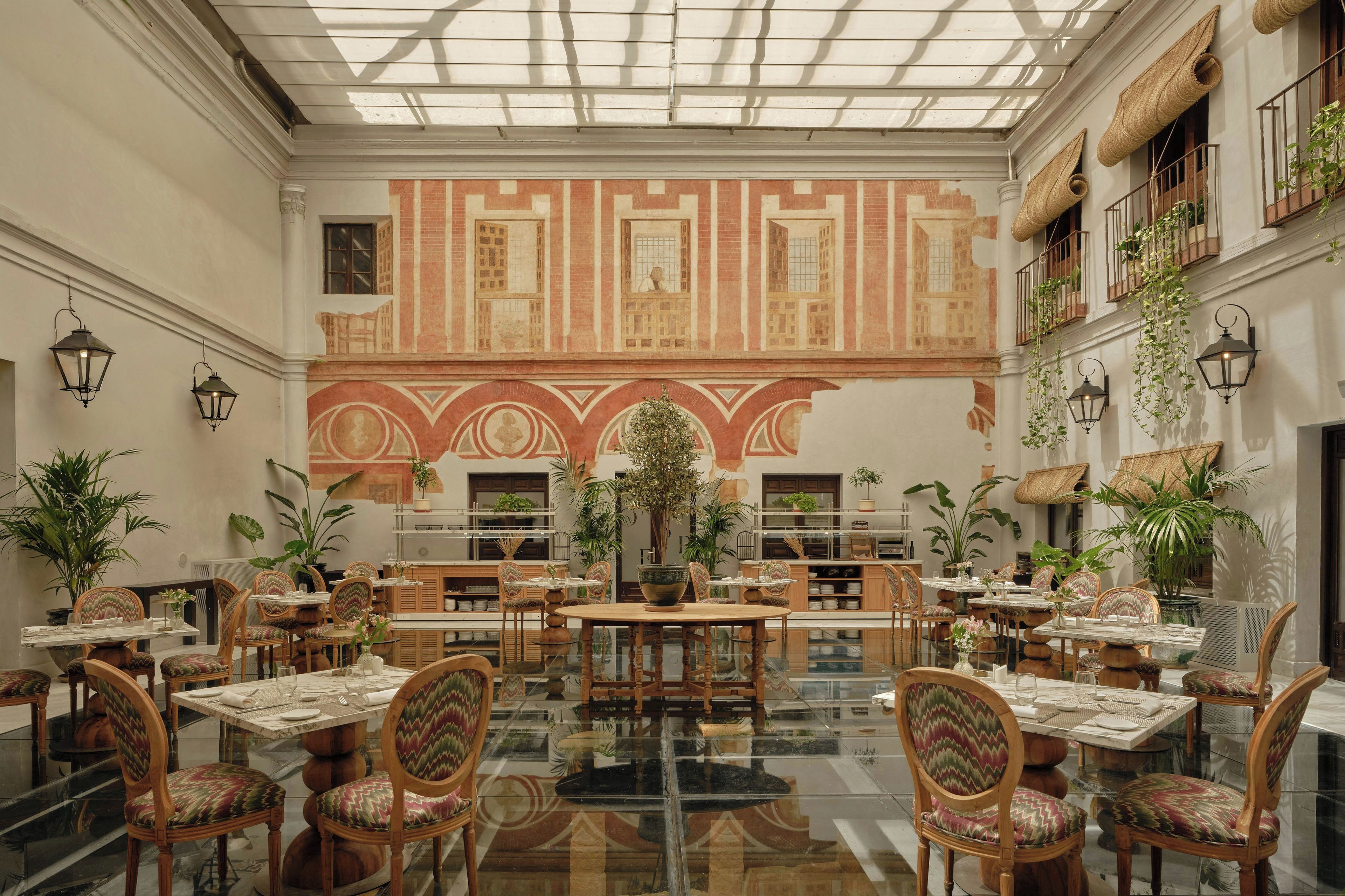 Un espacio interior amplio con varias mesas y sillas para comer, un mural grande en la pared y plantas en macetas.
