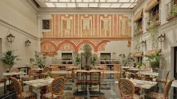 Un espacio interior amplio con varias mesas y sillas para comer, un mural grande en la pared y plantas en macetas.