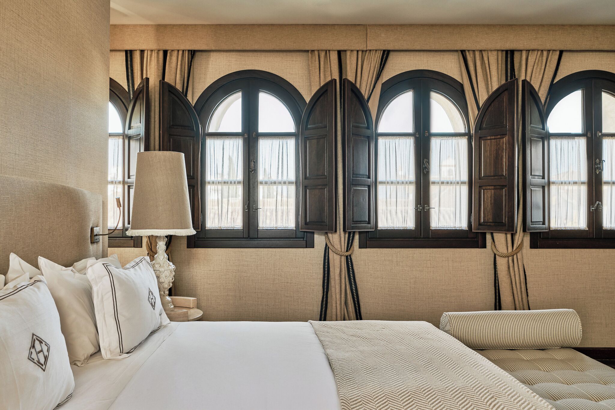 A hotel room with a large bed, a nightstand, a lamp, and three arched windows with curtains.