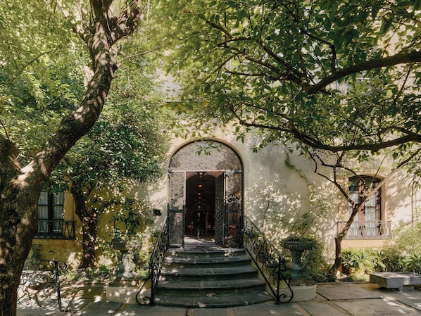 L’entrée d’un immeuble, avec un escalier menant à une porte en arc de cercle, le tout entouré d’arbres et de verdure.