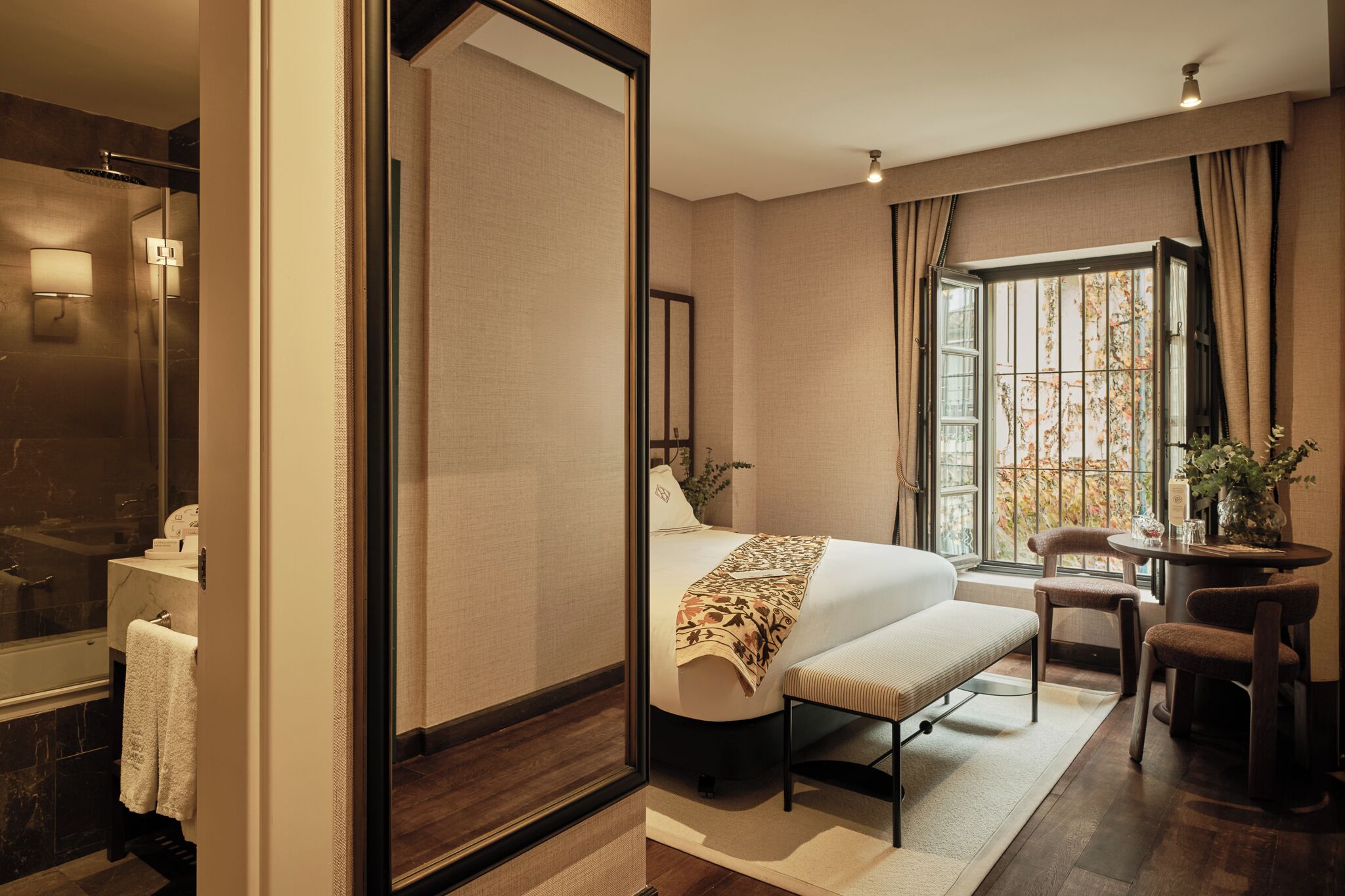 Une chambre d’hôtel avec un grand lit, un coin salon avec un banc, une petite table, une fenêtre avec des rideaux et une salle de bain avec une douche et un miroir.