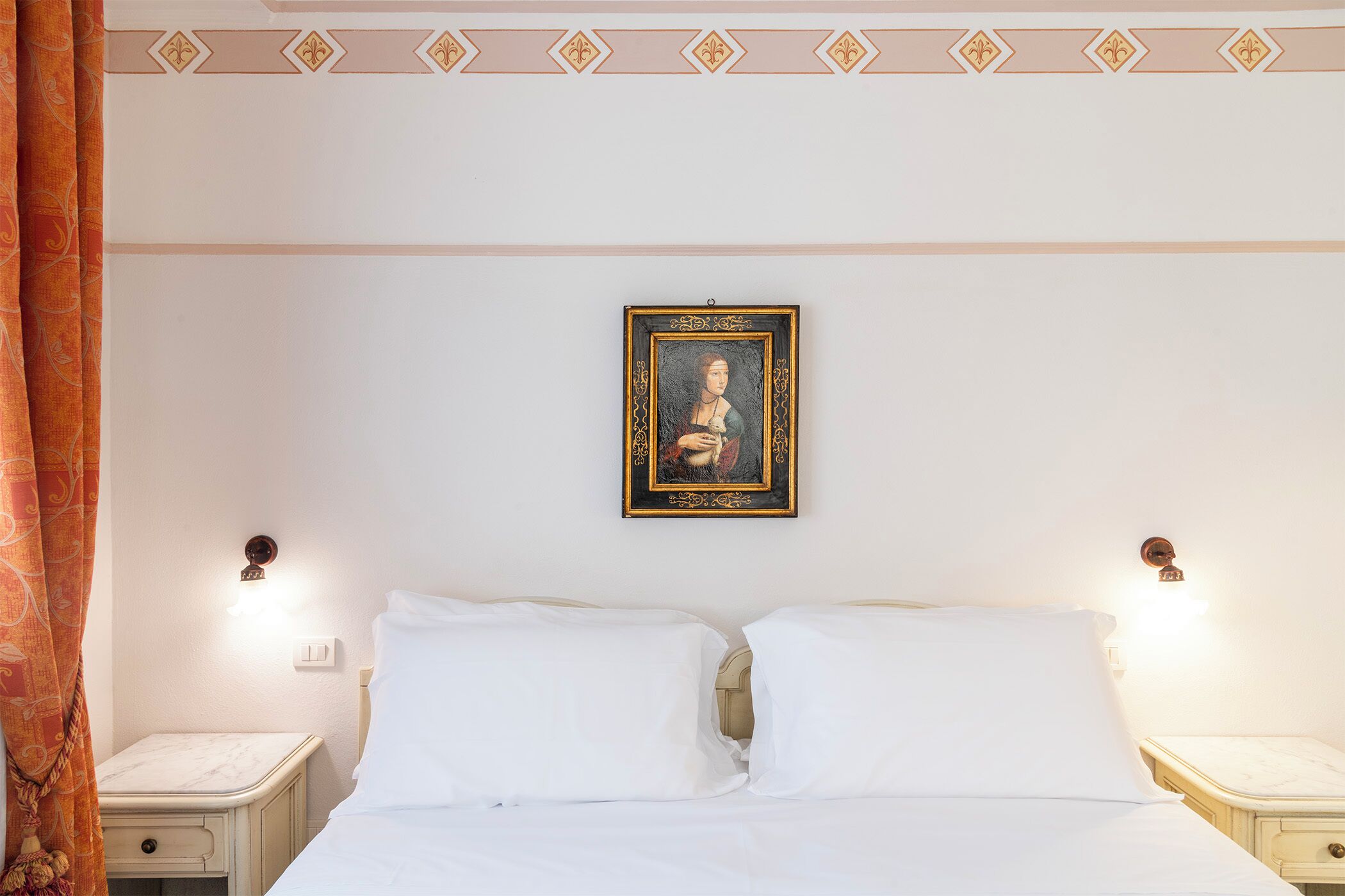 Une chambre d’hôtel comprenant un lit, deux tables de chevet, une petite table, un tableau accroché au mur et un motif décoratif au-dessus du lit.