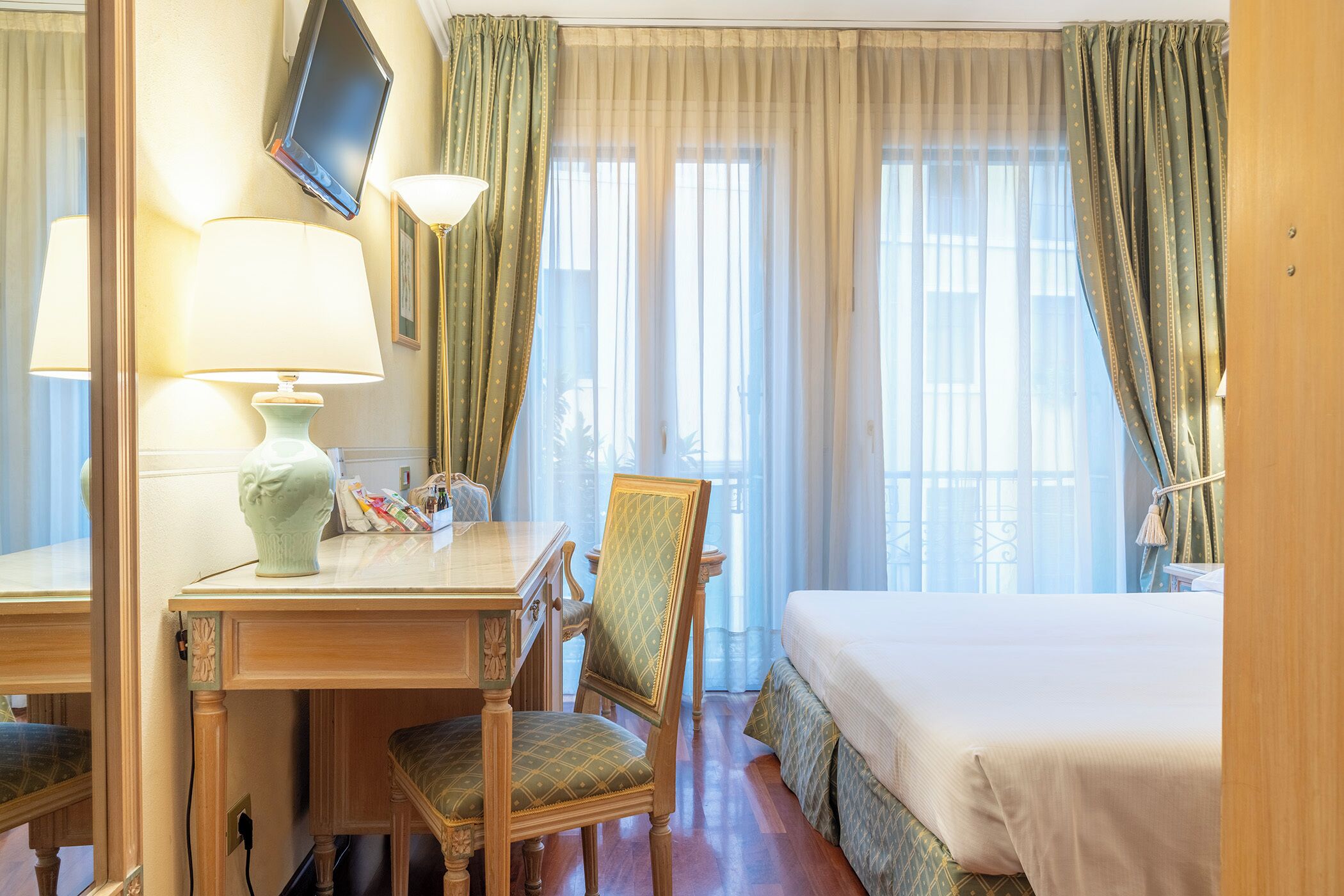 A hotel room with a bed, a desk with a lamp, a chair, and a television on the wall.