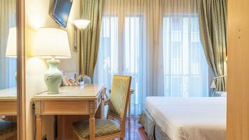 A hotel room with a bed, a desk with a lamp, a chair, and a television on the wall.