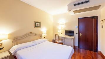 Habitación de hotel con cama, lámparas de noche, un escritorio con televisor, una puerta de madera y un cuadro en la pared.
