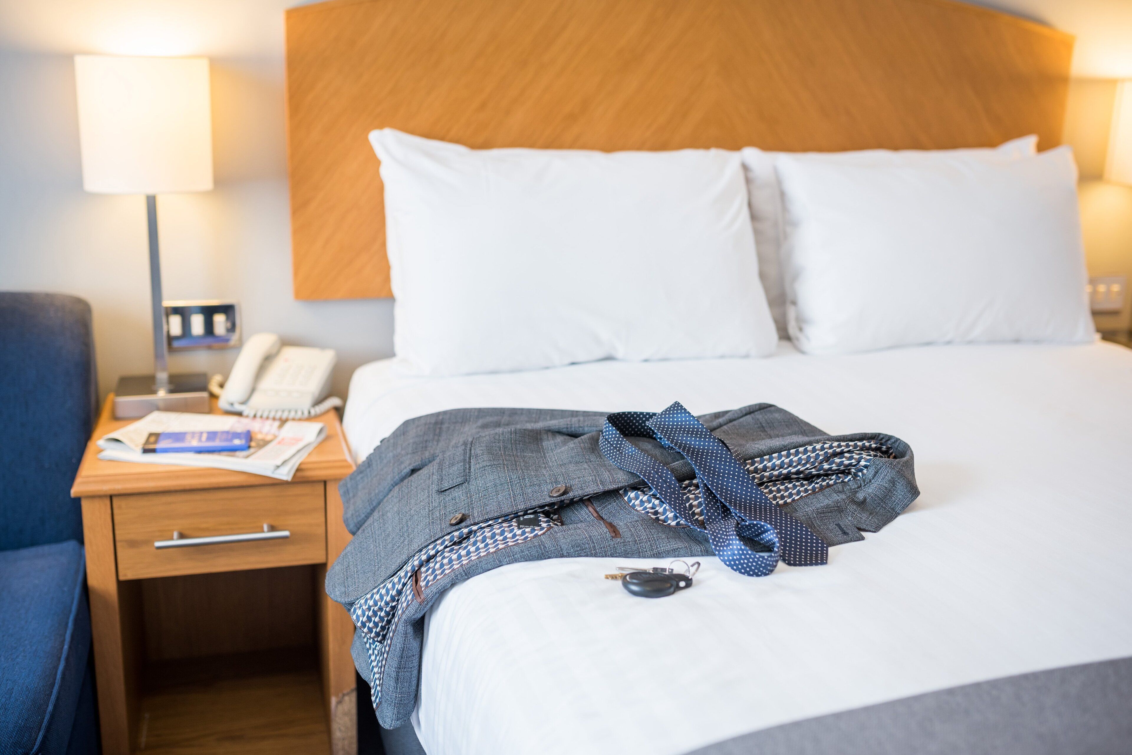 Una habitación de hotel con cama, mesita de noche con lámpara, teléfono y una silla azul.