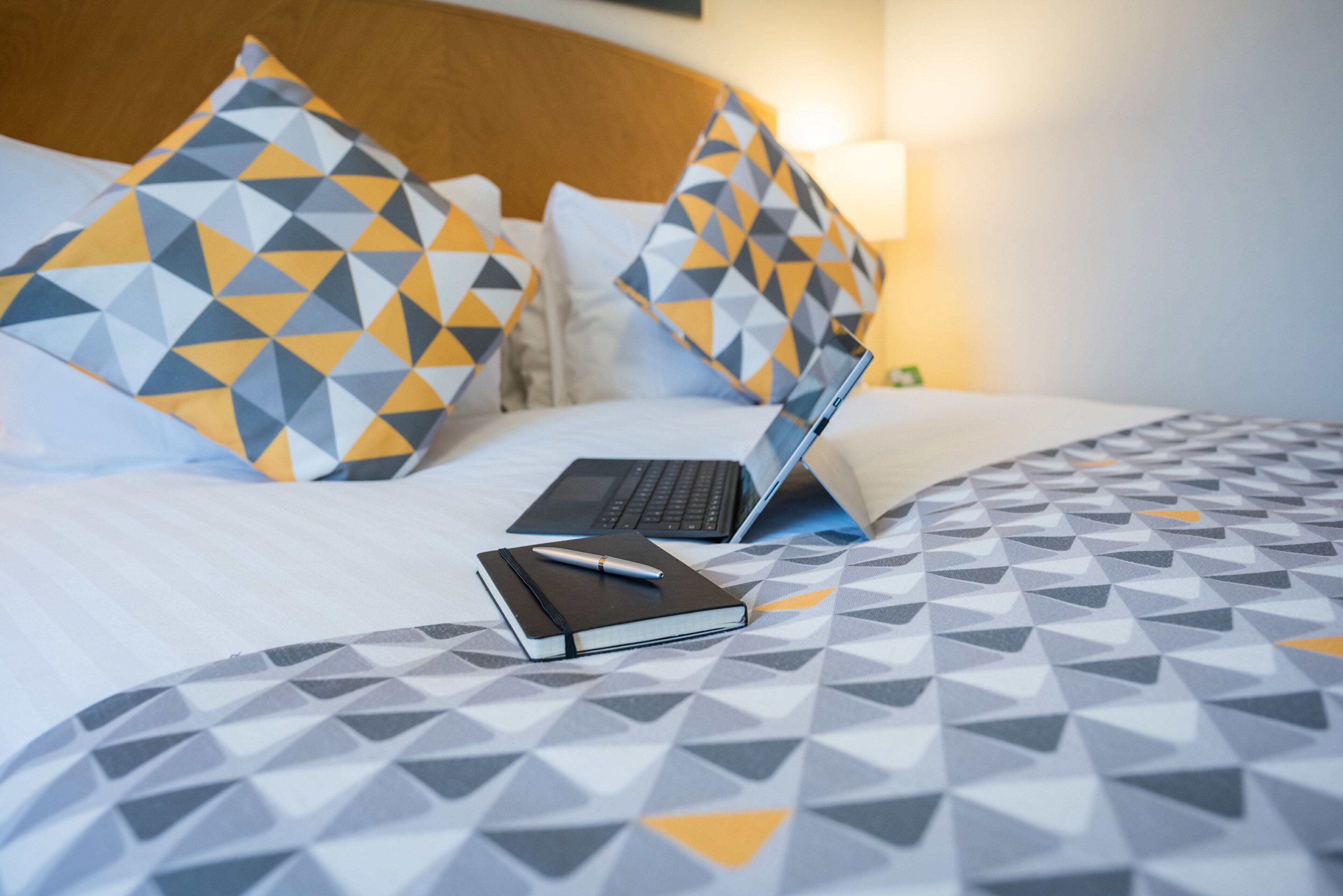 A bed with a laptop, a notebook, and a pen on a bed with a geometric patterned bedspread.