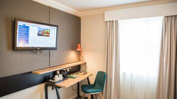 A hotel room with a desk, chair, TV, and a window with curtains.
