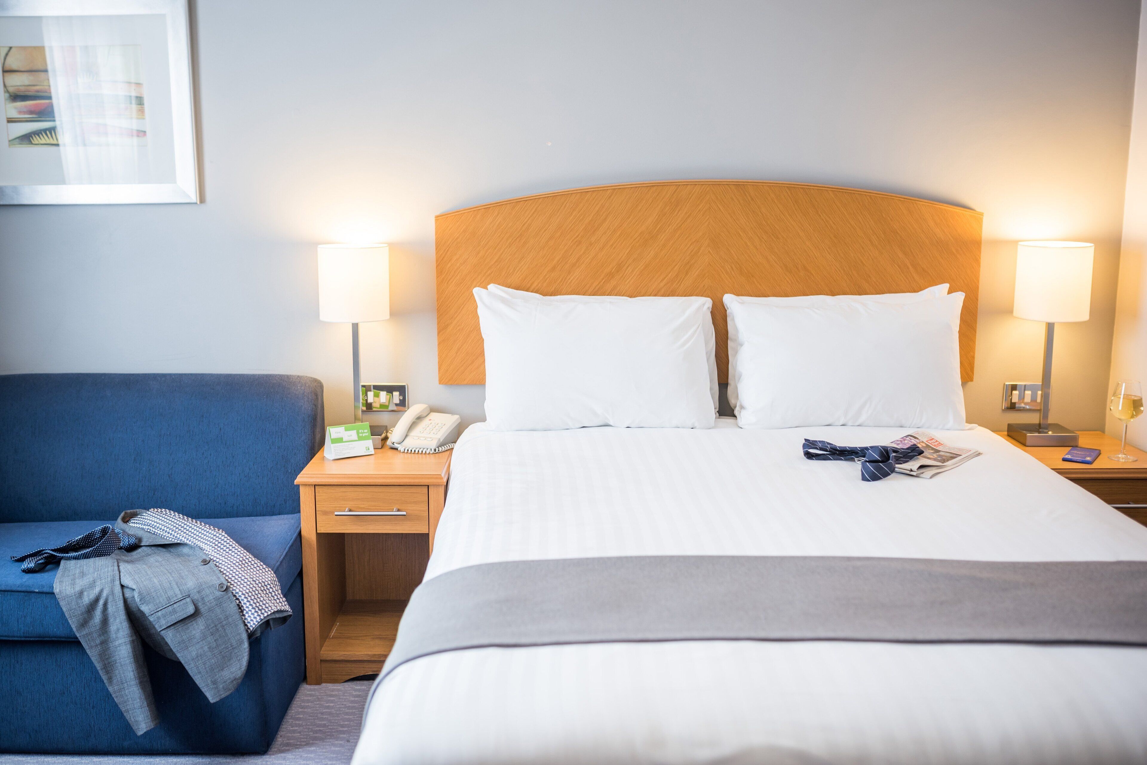 Hotellihuone, jossa on suuri sänky, kaksi yöpöytävalaisinta, puinen sängynpääty, sininen nojatuoli sekä yöpöytä, jossa on puhelin ja valaisin.