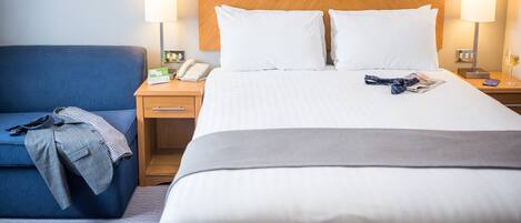 Hotellihuone, jossa on suuri sänky, kaksi yöpöytävalaisinta, puinen sängynpääty, sininen nojatuoli sekä yöpöytä, jossa on puhelin ja valaisin.