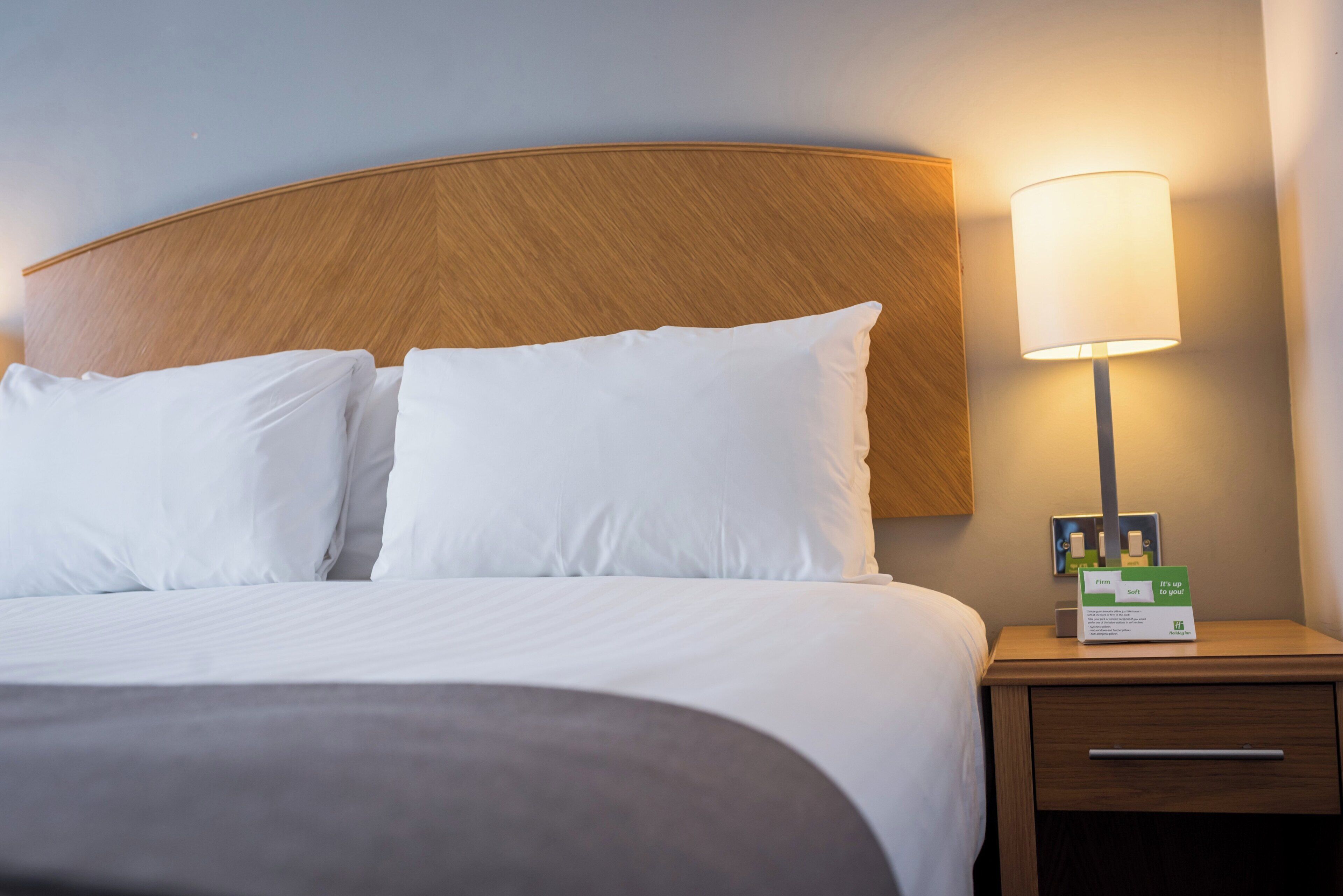 A hotel room with a bed, pillows, a nightstand, a lamp, and a card holder with a card.