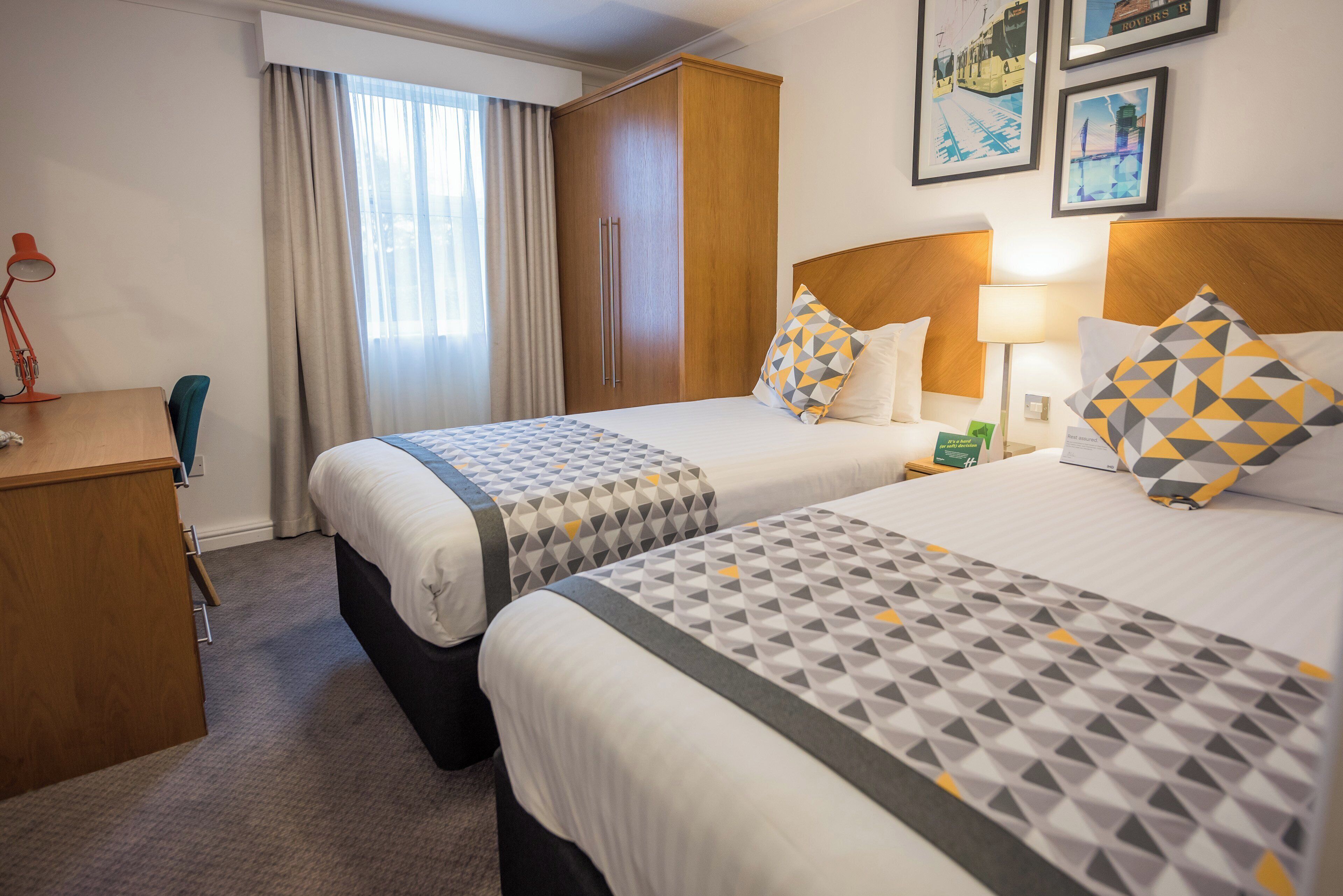 Ett hotellrum med två sängar, ett skrivbord, en stol, en lampa och ett fönster med gardiner.