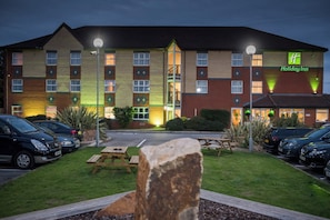 A Holiday Inn hotel with a parking lot, outdoor seating, and a stone feature in the foreground. - Holiday Inn Manchester West by IHG (Salford)