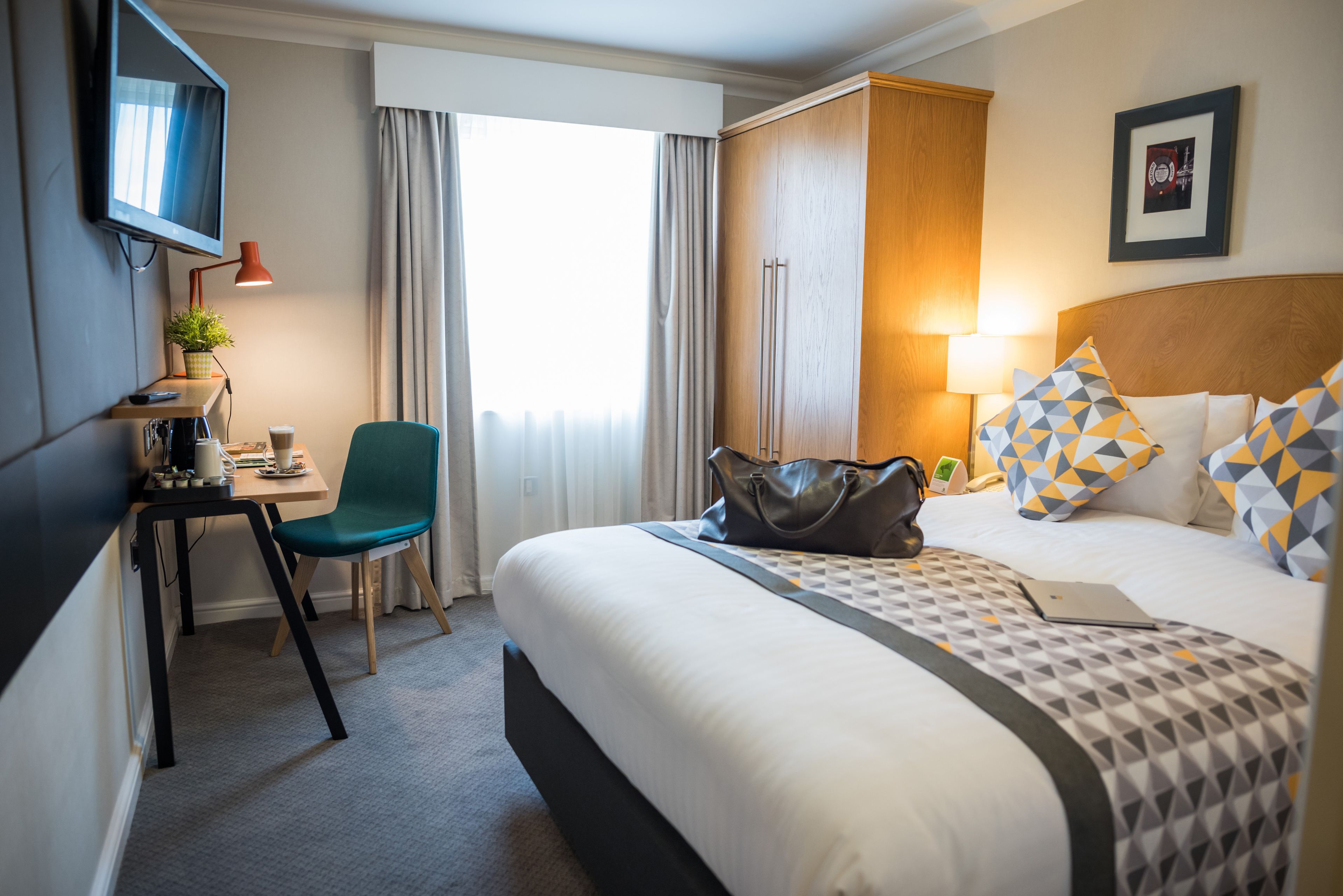 A hotel room with a bed, a desk with a chair, a TV, a lamp, and a window with curtains.