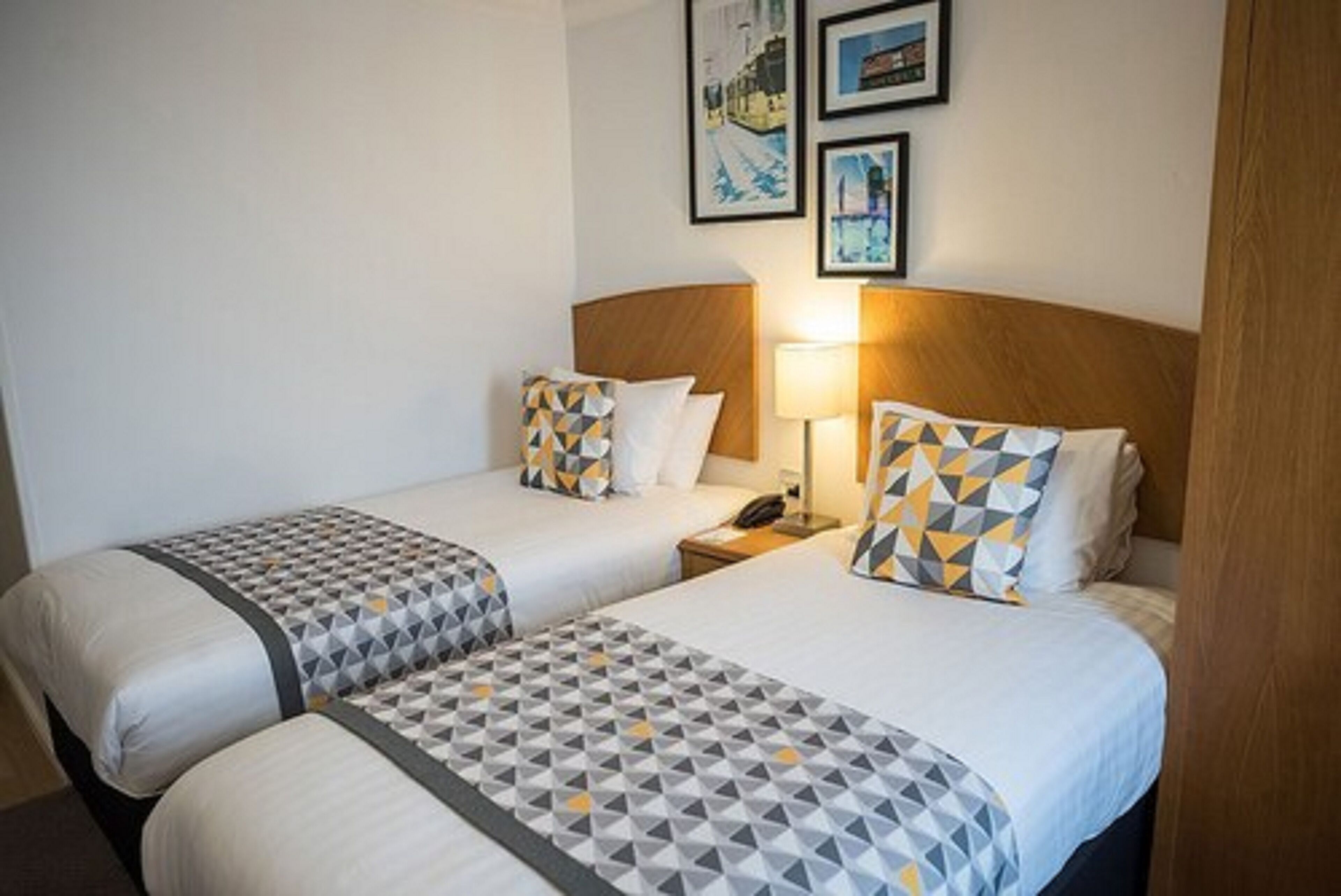 A hotel room with two beds, each with patterned bedding and pillows, a bedside table with a lamp, and framed pictures on the wall.