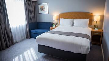 A hotel room with a large bed, a blue armchair, a bedside table with a lamp, and a window with curtains.