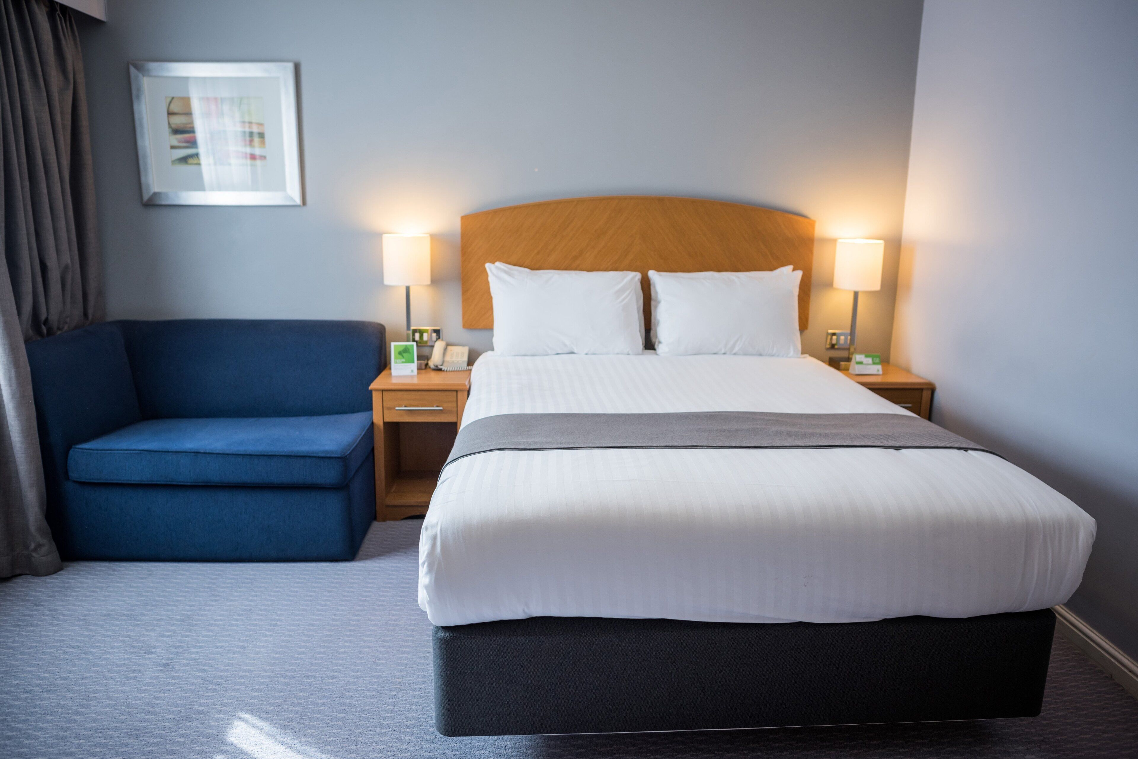 A hotel room with a large bed, a blue armchair, two bedside tables with lamps, and a framed picture on the wall.