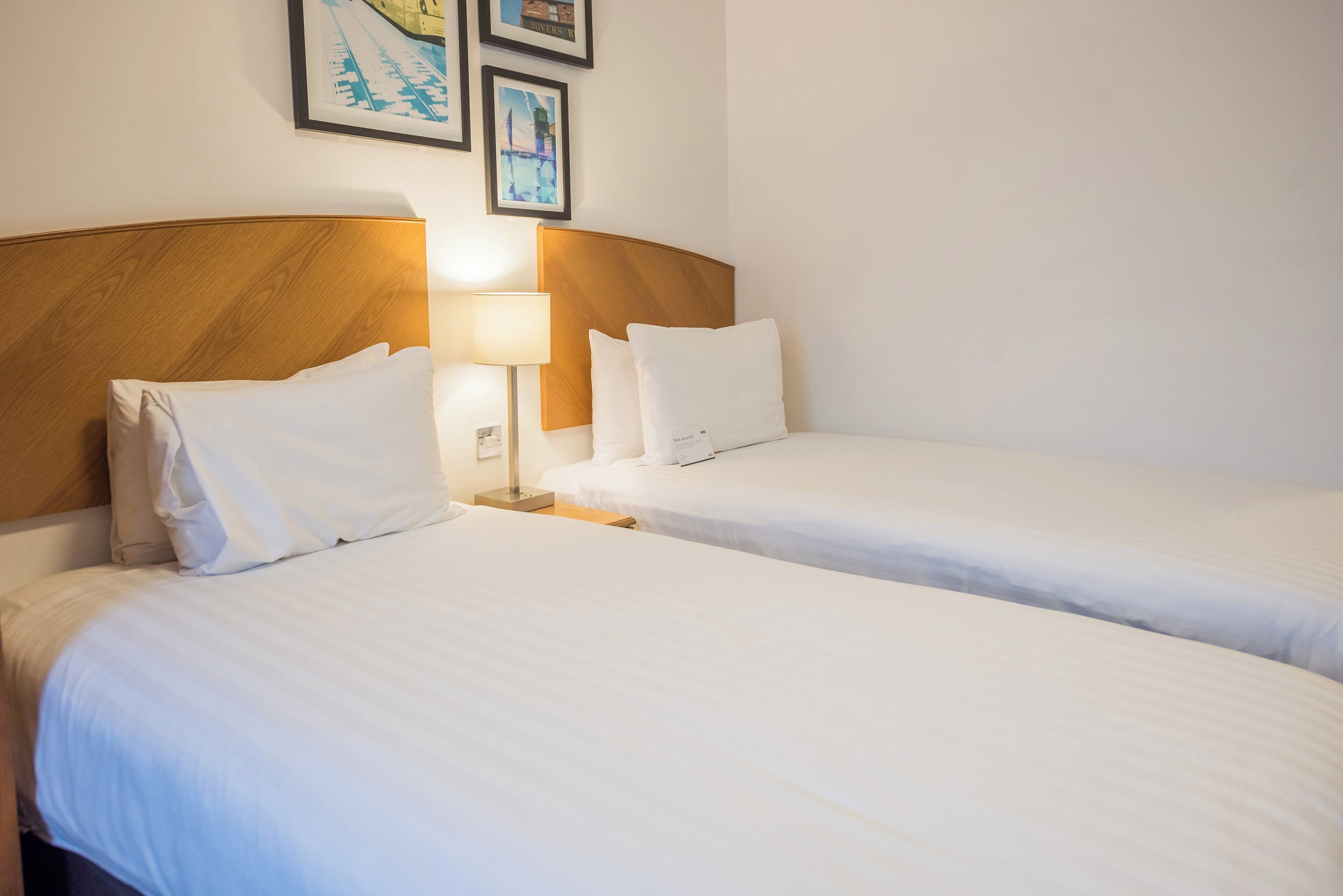 Habitación de hotel con dos camas individuales, cabecero de madera, mesita de noche con lámpara y cuadros en la pared.