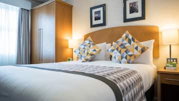 A hotel room with a large bed, two framed pictures on the wall, a wooden wardrobe, and a bedside table with a lamp.